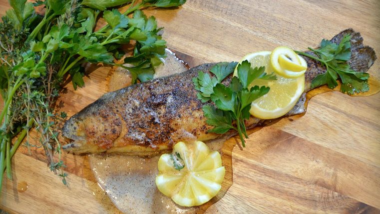 A grilled fish lies on a wooden board, surrounded by fresh herbs. Next to the fish are lemon slices and a decorative lemon blossom.