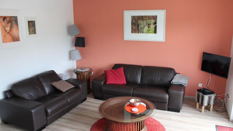 Un salon confortable avec deux canapés marron et une table basse ronde. Les murs sont partiellement peints en orange et quelques images sont accrochées de manière décorative au mur.