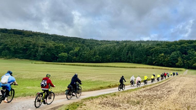 Four people are standing in front of a café and posing with an e-bike. They are smiling and wearing sporty clothing.
