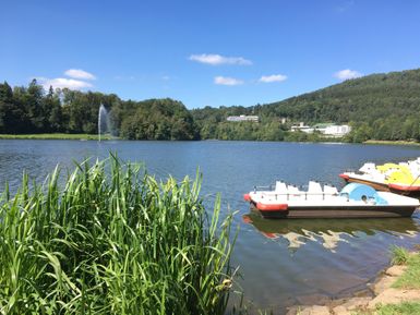 A calm lake surrounded by green shores and gentle hills. Some pedal boats can be seen at the water, and the sky is clear and blue.