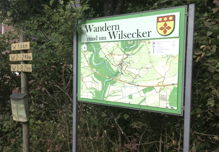 Infotafel in Wilsecker zeigt Wanderwege und eine Karte. Daneben ein Wegweiser mit Entfernungen. Umgeben von B&auml;umen und Str&auml;uchern., &copy; Tourist-Info Bitburger Land M.Mayer