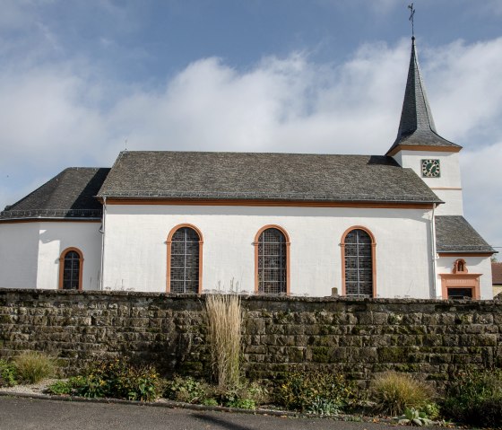 L'église paroissiale Saint-Martin à Dockendorf avec sa tour d'horloge, entourée d'un mur de pierre et d'un jardin. Le ciel est légèrement nuageux., © TI Bitburger Land