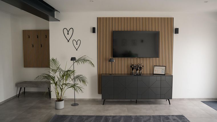 Ein modernes Wohnzimmer mit einer Holzvertäfelung und einem Fernseher an der Wand. Eine Pflanze und ein schwarzes Sideboard ergänzen die schlichte, elegante Einrichtung.