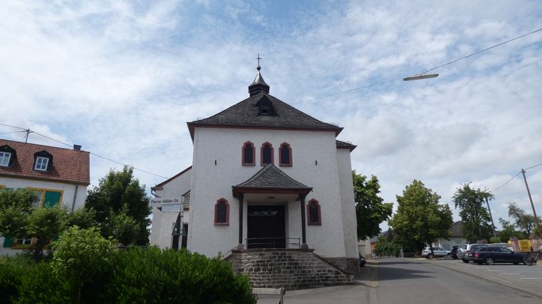 Frontansicht einer Kirche mit weißer Fassade und rotem Fensterrahmen, umgeben von Bäumen und geparkten Autos.