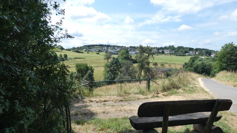 Landschaft mit Bank, Feldweg und Dorf im Hintergrund.
