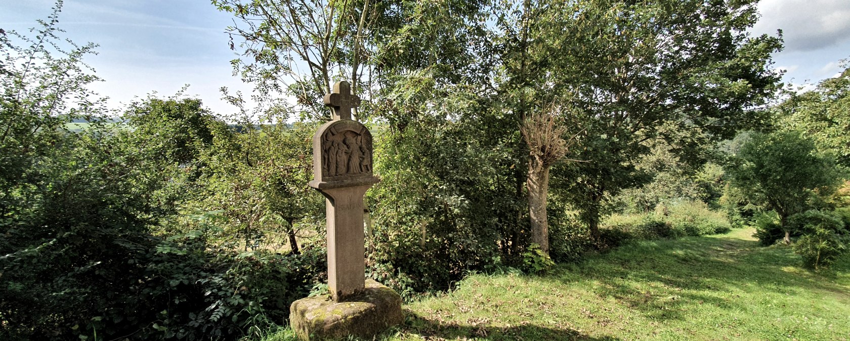 Ein steinernes Kreuz steht auf einer grünen Wiese, umgeben von Bäumen und Sträuchern. Der Himmel ist blau mit wenigen Wolken., © Tourist-Information Bitburger Land