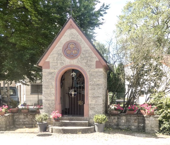 Small chapel in Idenheim, surrounded by flowers and trees. A street sign shows 'Meilbrücker Straße'. The chapel has a pointed roof and a lattice gate., © TI Bitburger Land