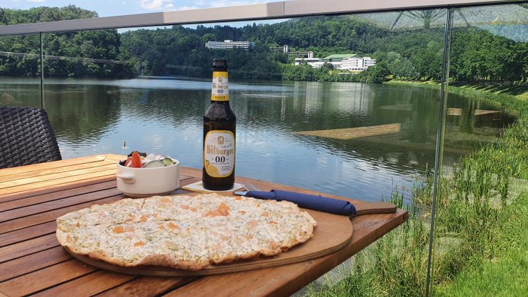 Ein Tisch mit einer großen Pizza, einer Flasche Bier und einem kleinen Salat. Im Hintergrund ist ein malerischer See und grüne Landschaft zu sehen.