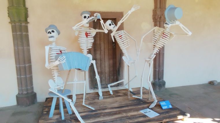 Vier fantasievolle Skelettfiguren, die Musikinstrumente spielen und tanzen. Sie stehen auf einer Holzplattform in einem hellen Raum.