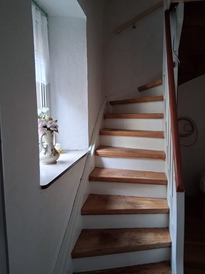 Ein heller Treppenaufgang mit Holzstufen. Ein Fenster mit Blumen schmückt die Wand.