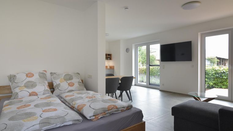 Ein modernes Wohn- und Schlafzimmer mit einem Doppelbett, einem Tisch und Stühlen. Große Fenster bieten viel Licht und einen Blick ins Grüne.