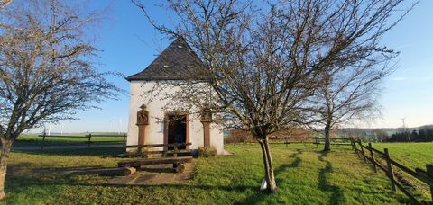 Kleine Kapelle auf einer Wiese mit Bäumen und Windrädern im Hintergrund.
