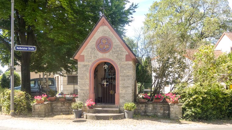 Kleine kapel in Idenheim met bloemen en bomen op de achtergrond.
