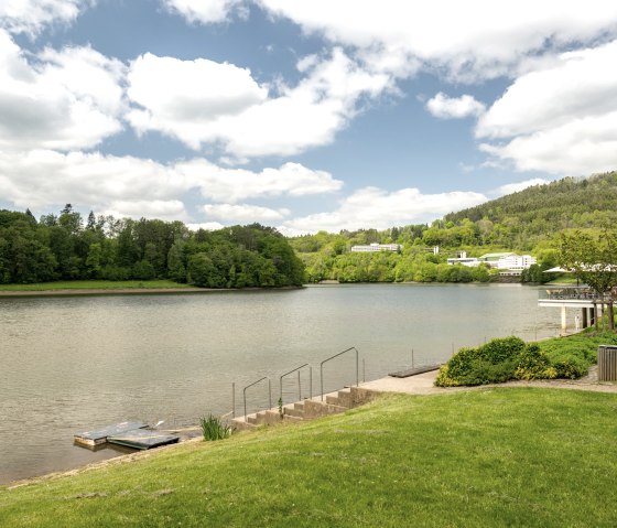 Het Bitburg stuwmeer in Biersdorf am See, omgeven door groene bomen en heuvels, onder een bewolkte hemel. Een loopbrug leidt naar het water., © Eifel Tourismus GmbH, Dominik Ketz