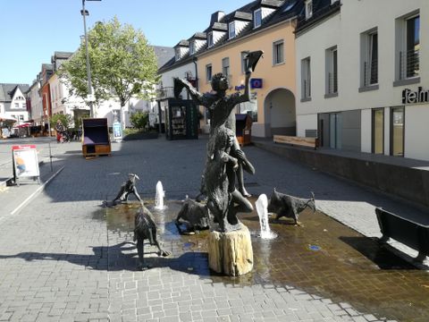 Eine Statue mit einer Frau und mehreren Ziegen in einem Brunnen auf einer gepflasterten Straße. Umgeben von modernen Gebäuden und Bäumen.
