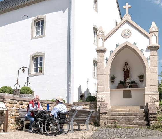 Deux hommes, dont l'un en fauteuil roulant, sont assis sur un banc devant un bâtiment blanc abritant une statue religieuse dans une niche. Journée ensoleillée., © Naturpark Südeifel, Thomas Urbany