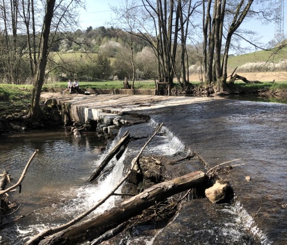 Twee mensen zitten op een brug over de Nims in een groene, landelijke omgeving. De rivier stroomt rustig onder de brug door., © Tourist-Information Bitburger Land_Uschi Hallet