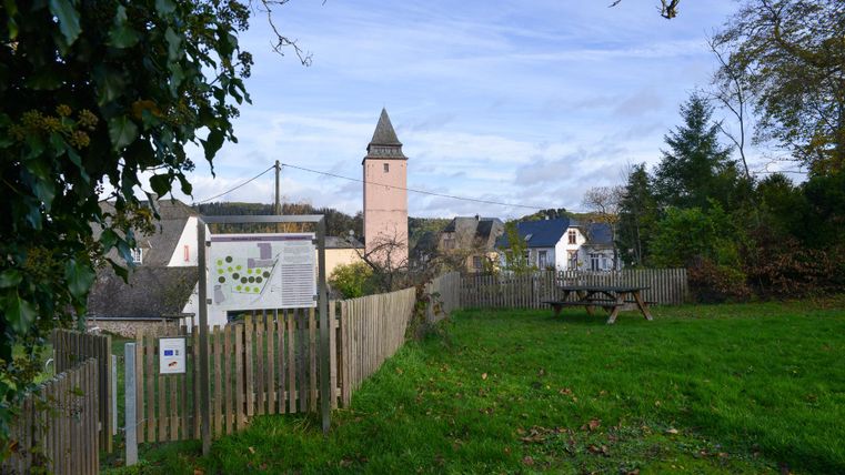 Eine ruhige Szene mit einem kleinen Garten und einem Holzzaun. Im Hintergrund ist ein historischer Turm und einige Häuser vor einem klaren Himmel zu sehen.