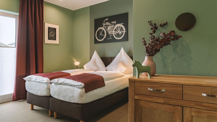 Ein einladendes Schlafzimmer mit zwei Betten und einer grünen Wandfarbe. Der Raum enthält moderne Möbel und dekorative Elemente wie ein Fahrradbild und einen Holztisch.
