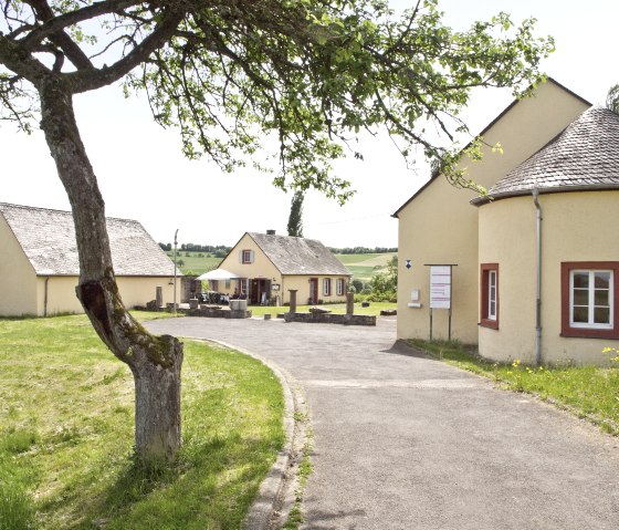 Gelbe Gebäude der Römischen Villa Ortrang in Fließem, umgeben von grüner Landschaft und einem Baum im Vordergrund., © Tourist Information Bitburger Land