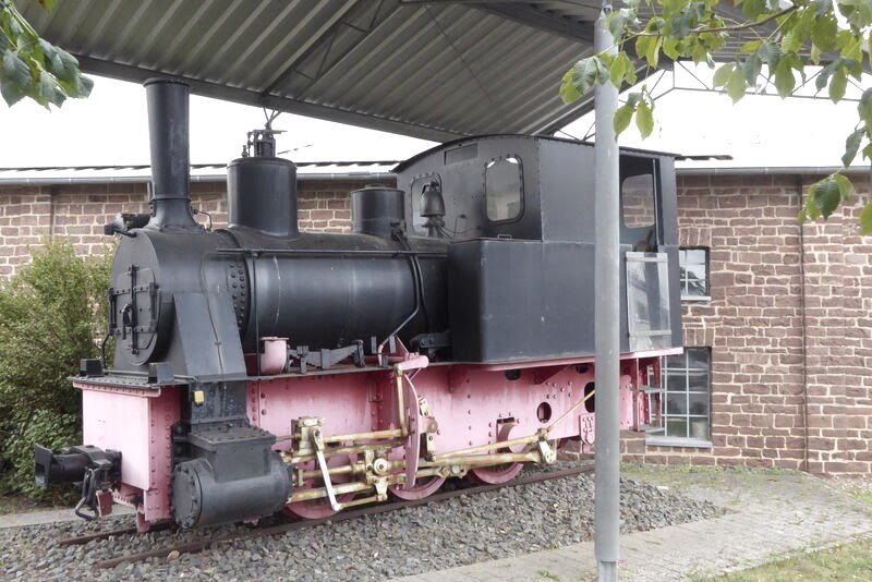 Eine alte Dampflokomotive steht unter einem Schutzdach vor einem Backsteingebäude. Die Lokomotive ist schwarz und rosa gestrichen., © Eifelverein Ortsgruppe Speicher