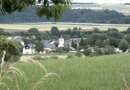 Ein Dorf mit einer Kirche in der Mitte, umgeben von grünen Feldern und Bäumen, unter einem bewölkten Himmel., © TI Bitburger Land