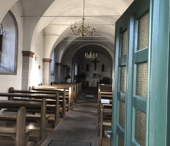 View into a small chapel with wooden benches, an altar and chandeliers. The green door is open, light falls through the windows., &copy; TI Bitburger Land - Steffi Wagner