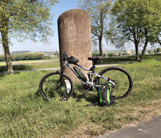Een fiets leunt tegen een stenen pilaar op een groene weide. Op de achtergrond zijn bomen en een uitgestrekt landschap te zien., © TI Bitburger Land, Steffi Wagner