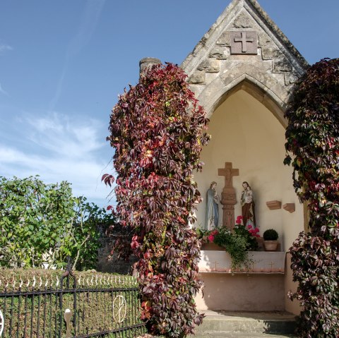 Een kleine kapel in Wolsfeld, omgeven door rode klimplanten. Binnen zijn religieuze beelden te zien. De lucht is helder en blauw., © Tourist-Information Bitburger Land