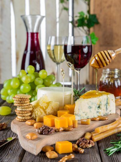 Un plateau élégant avec différents types de fromage, du miel et des noix. En arrière-plan, on peut voir des verres à vin et des raisins.