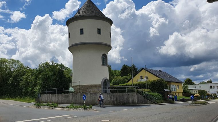 Ein runder Wasserturm mit spitzem Dach steht neben einer Straße, umgeben von Bäumen und Häusern. Ein Radfahrer fährt vorbei, der Himmel ist bewölkt.