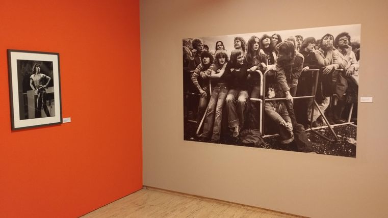 An exhibition space with two pictures on the wall. On the left, a black-and-white photo of a person, on the right, a large black-and-white image of a group of young people.