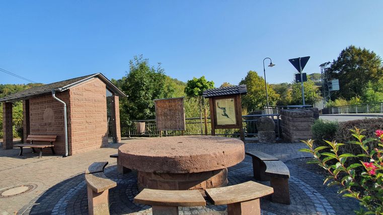 Dorfplatz in Brecht mit Rundbank und Infotafeln, umgeben von Bäumen und einem kleinen Gebäude.
