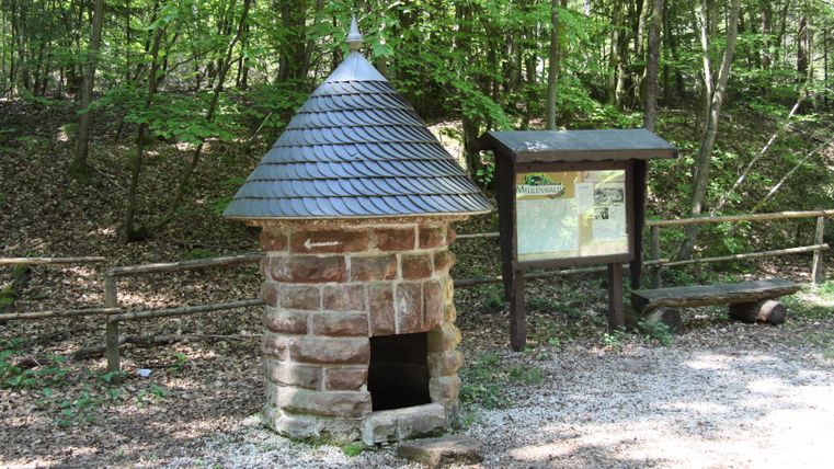 Stone fountain with pointed roof in the forest, next to it an information board and a bench.
