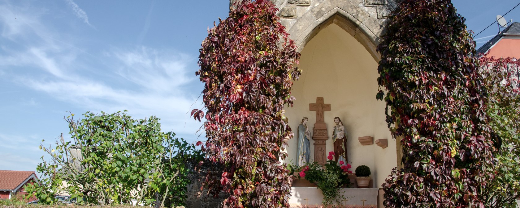 Een kleine kapel in Wolsfeld, omgeven door rode klimplanten. Binnen zijn religieuze beelden te zien. De lucht is helder en blauw., © Tourist-Information Bitburger Land