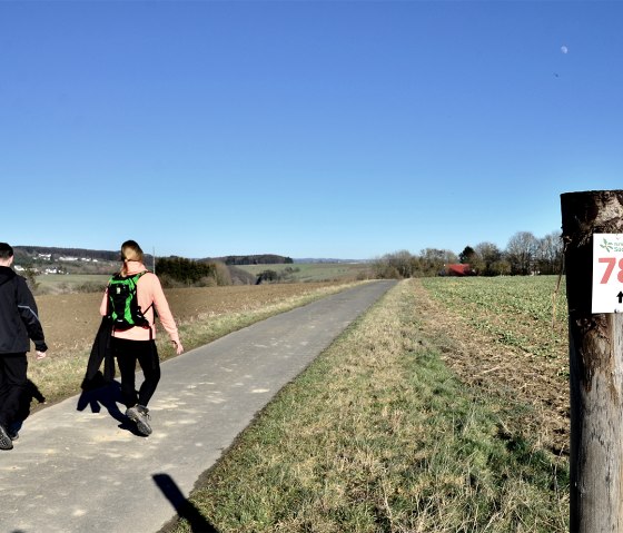 Two hikers on a path in the Southern Eifel Nature Park. A post with the number 78 stands at the edge of the path. The sky is clear and blue., © TI Bitburger Land