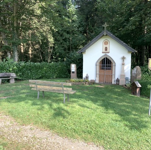 Eine kleine Kapelle im Wald mit einer Bank und einer Infotafel davor. Die Umgebung ist grün und friedlich., © TI Bitburger Land