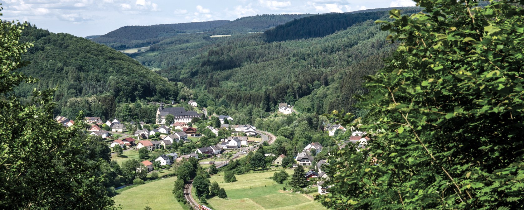 Panoramablick auf ein Dorf inmitten grüner Hügel und Wälder. Ein Zug fährt auf einer Bahnstrecke durch die Landschaft., © TI Bitburger Land