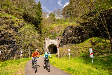 Twee fietsers genieten van een pittoresk landschap op een fietspad. Op de achtergrond is een oude tunnel en groene heuvels te zien.