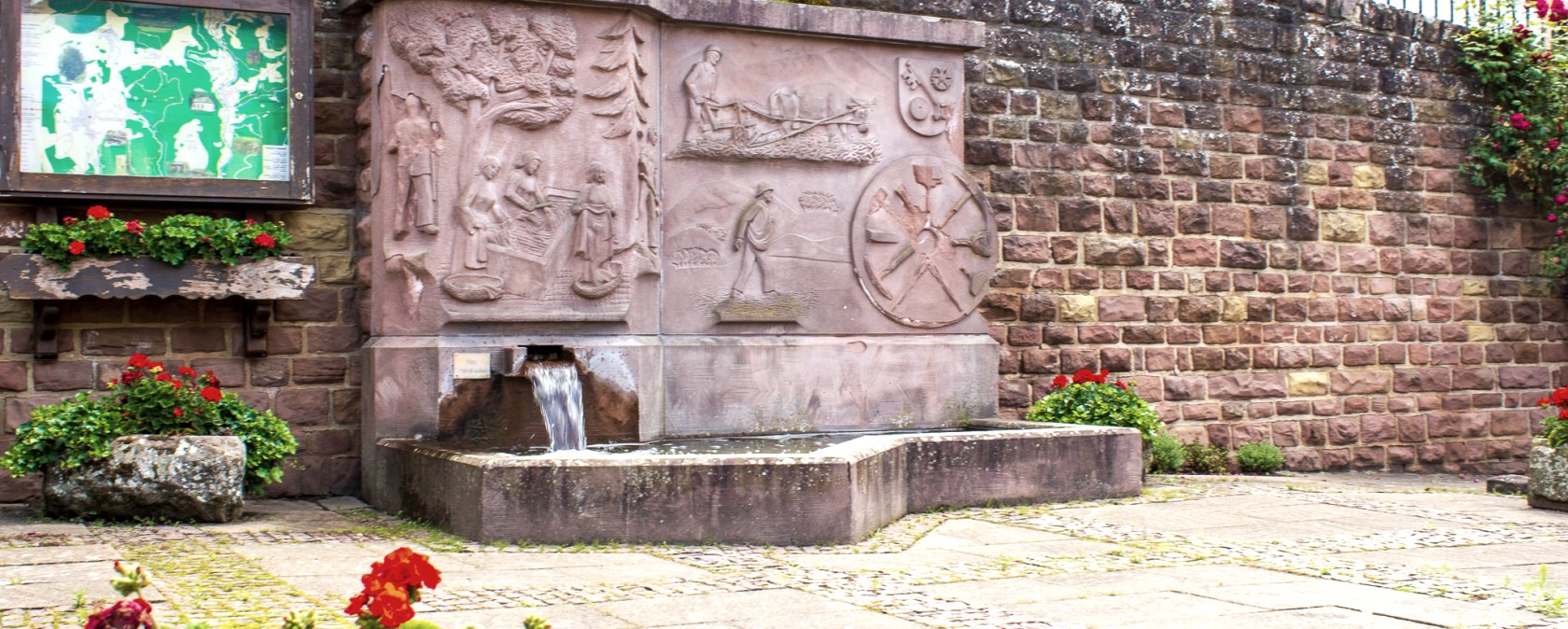 Ein steinerner Dorfbrunnen mit Reliefs und fließendem Wasser, umgeben von roten Blumen und einer Infotafel an einer Steinmauer., © TI Bitburger Land - M. Mayer