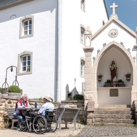 Twee mannen, één in een rolstoel, zitten op een bankje voor een wit gebouw met een religieus standbeeld in een nis. Zonnige dag., © Naturpark Südeifel, Thomas Urbany