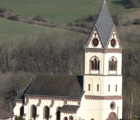 A church with a high tower stands in a green, rural landscape. The tower has a pointed roof and decorative windows., © TI Bitburger Land