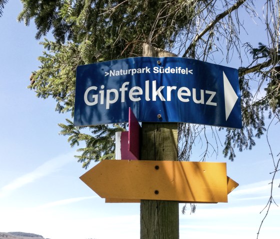 Ein Wegweiser im Naturpark Südeifel zeigt zum Gipfelkreuz. Der blaue Himmel und grüne Zweige rahmen das Schild ein., © TI Bitburger Land