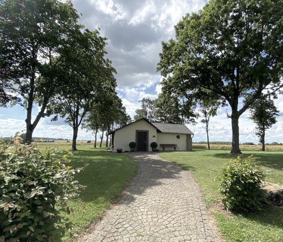 Un chemin pav&eacute; m&egrave;ne &agrave; un petit b&acirc;timent entour&eacute; d'arbres et de prairies vertes. Le ciel est nuageux., &copy; B. Milbach