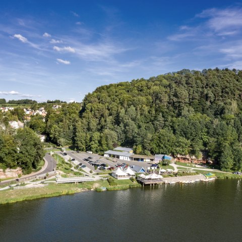 Vue aérienne du lac de barrage de Bitburg avec la forêt adjacente, les bâtiments et les parkings. Le ciel est clair et bleu., © team360 B. Treib