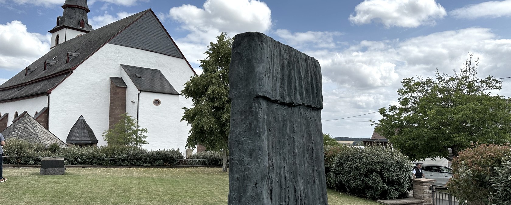 Kerk met spitse toren, modern beeldhouwwerk op de voorgrond, groen gazon, bomen en blauwe lucht met wolken.
