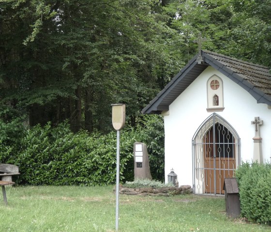 A small white chapel with a cross and barred door stands in the greenery. Next to it is a bench and a sign. Surrounded by trees and bushes., © TI Bitburger Land