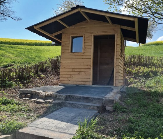 Petite maison en bois sur une colline avec un chemin pavé et un escalier, entourée d'une prairie verte et d'arbres sous un ciel bleu., © A. Girards