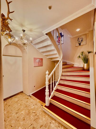 An elegant staircase with a white railing and red carpet. Pictures hang on the walls, and a plant arrangement creates a cozy atmosphere.