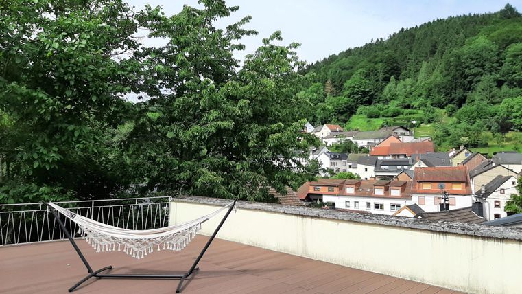 Une terrasse lumineuse avec un hamac et vue sur le village et les arbres environnants. Le paysage est vert et accueillant.